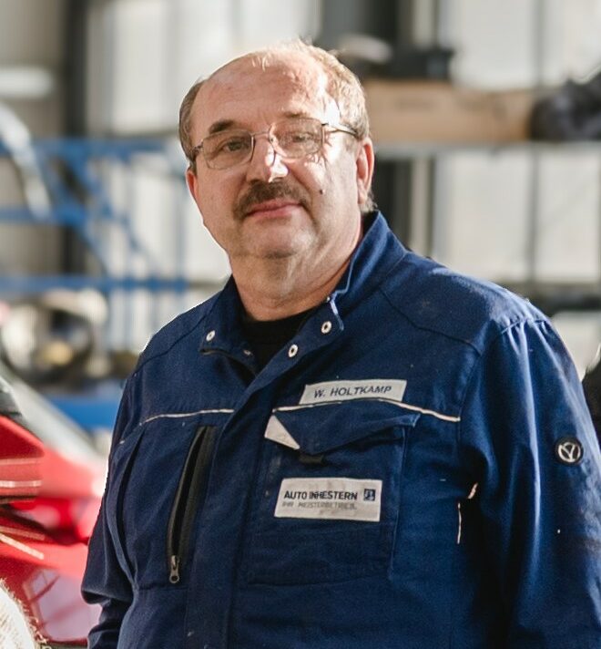 Person in blauer Arbeitskleidung mit Namensschild „W. Holtkamp“ und Logo von „Auto Inhestern“, im Hintergrund eine Werkstattumgebung.